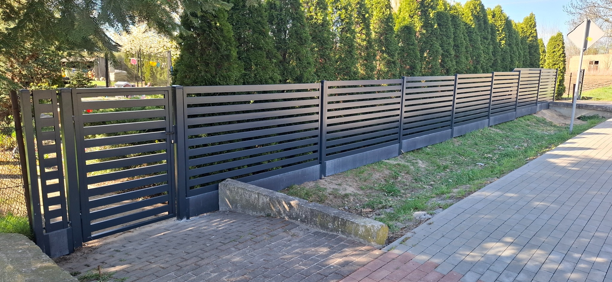 Nowoczesne, poziome, szare przęsła z metalu tworzące bramę i ogrodzenie na tle zielonych tuj i kostki brukowej.