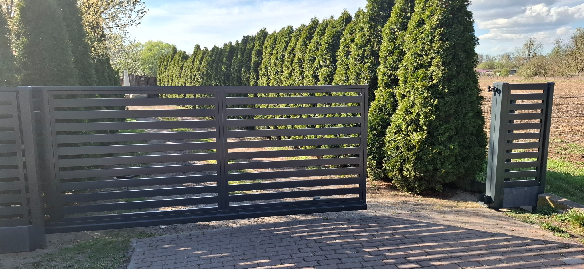 Automatyczna brama wjazdowa przesuwna z poziomych profili, otwierająca się na posesję z rzędem wysokich tuj, widoczny napęd bramy.