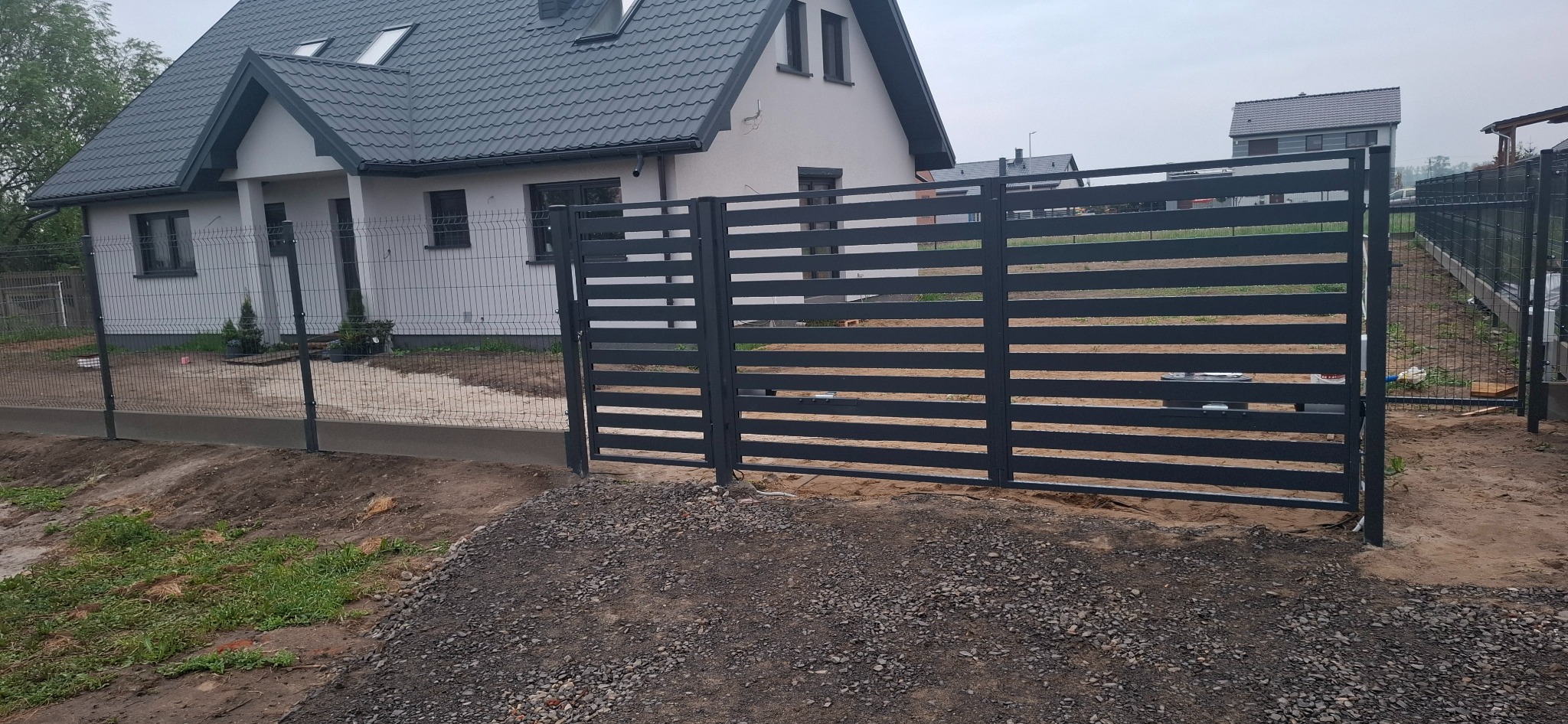 Nowoczesna brama przesuwna z poziomych profili, kolor antracyt, w Olszance. Widoczny fragment ogrodzenia panelowego i domu w tle. Minimalistyczny design.
