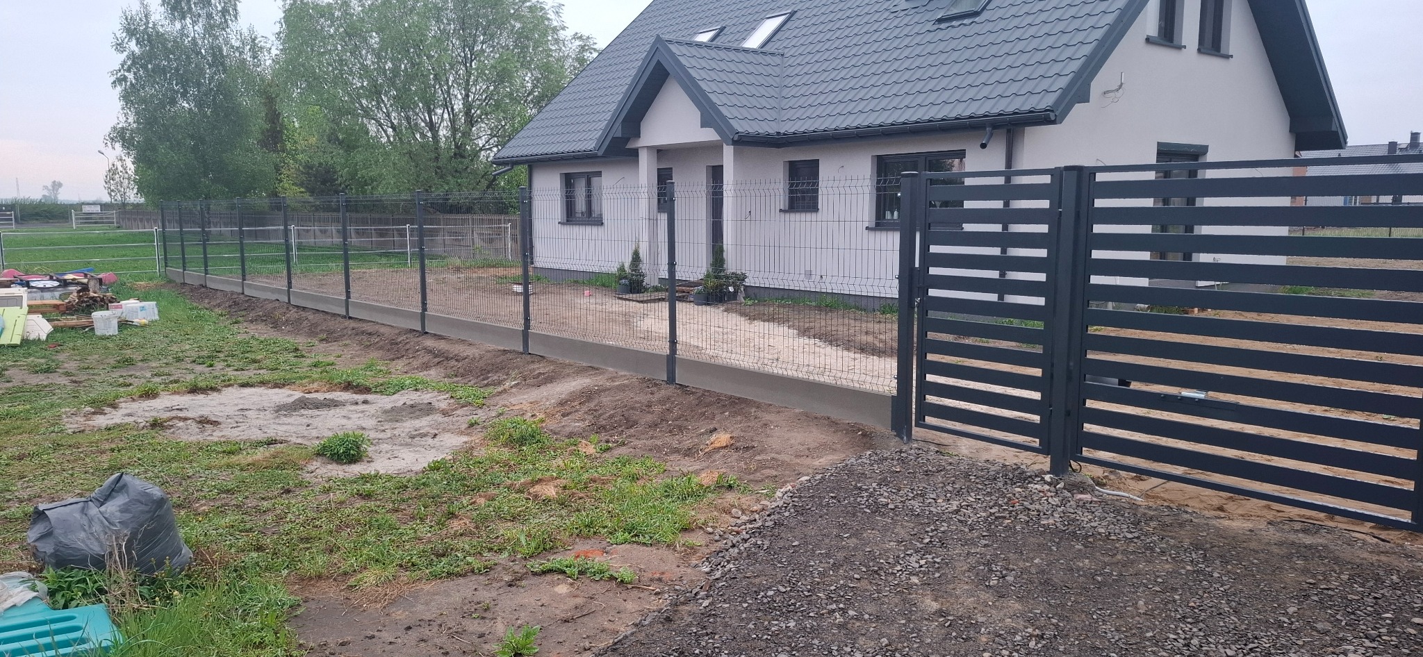 Nowoczesne ogrodzenie panelowe z bramą wjazdową, szara kolorystyka, betonowa podmurówka, widok na dom jednorodzinny w tle.