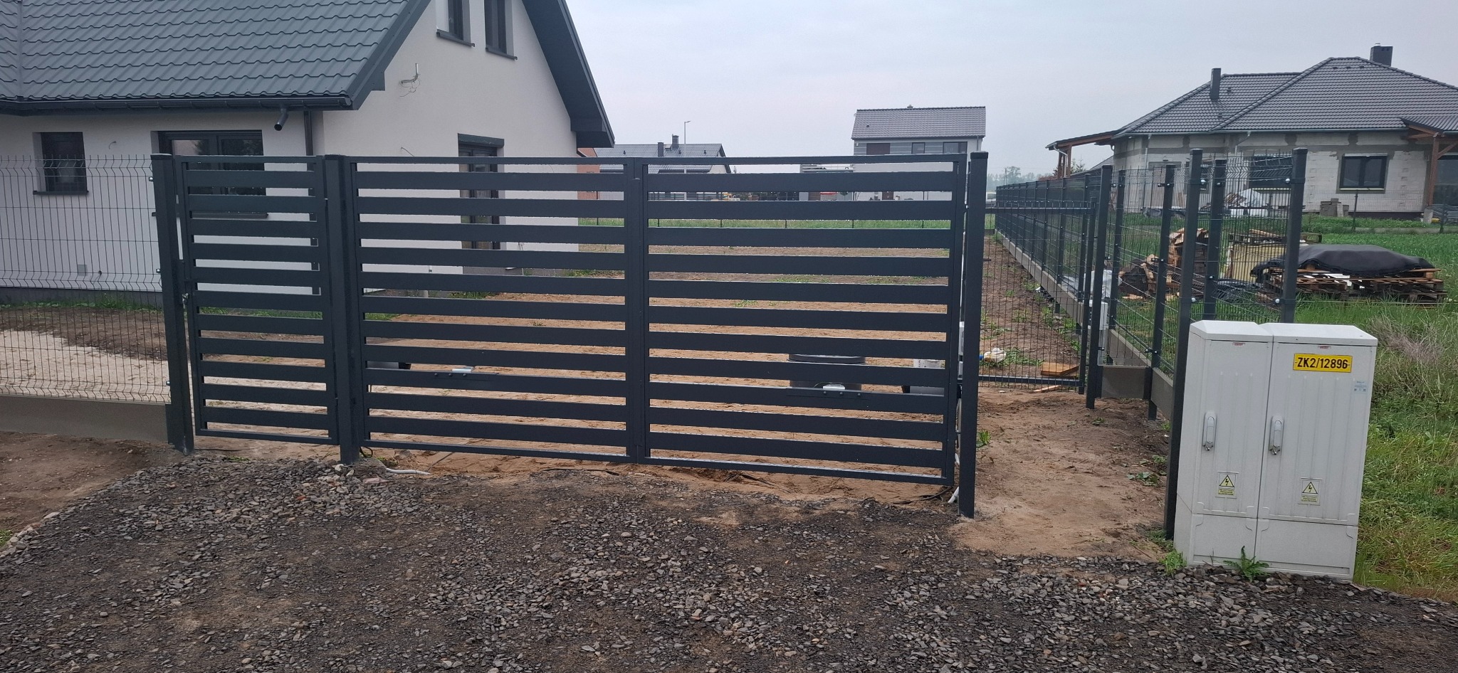 Nowoczesna, pozioma brama wjazdowa z metalowymi panelami w kolorze grafitowym. Widoczna automatyka bramy. W tle domy jednorodzinne i siatka ogrodzeniowa.