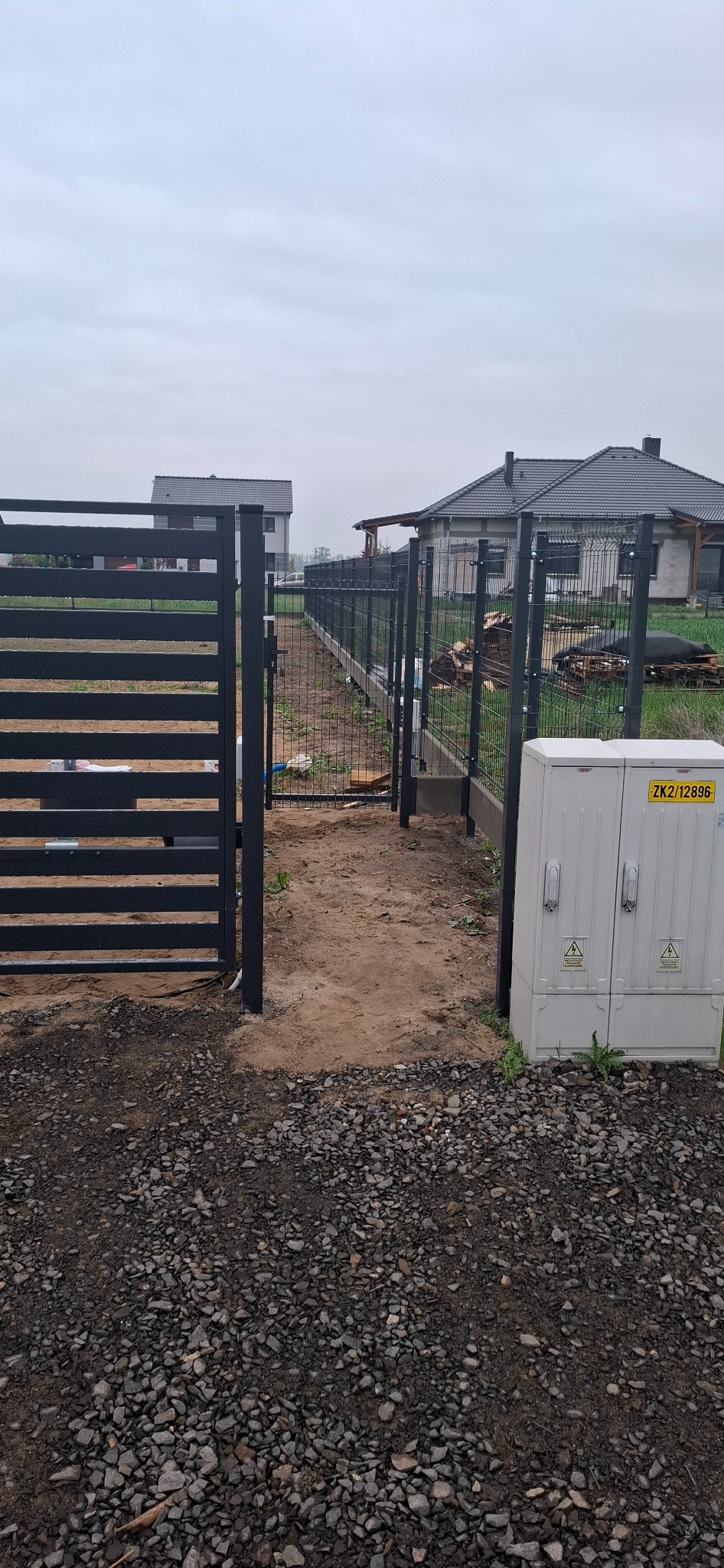 Nowoczesne ogrodzenie panelowe w kolorze grafitowym, z elementami siatki. Widok na posesję z ulicy, w tle domy jednorodzinne. Słupki wbetonowane, teren wyrównany.