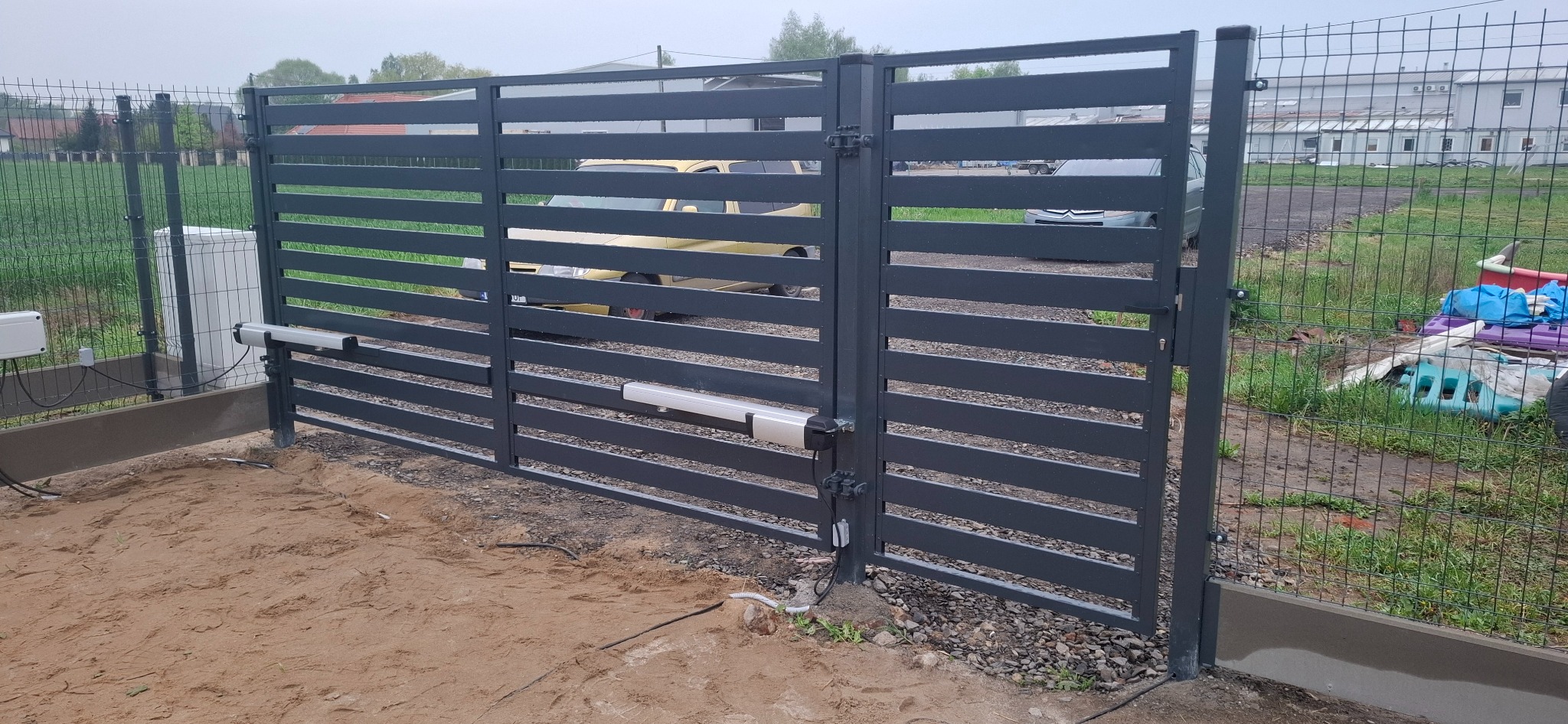 Nowoczesna, automatyczna brama metalowa z poziomymi szczeblami, szara, zamontowana przy ogrodzeniu z siatki. Widoczne elementy automatyki bramy i podłączenia elektryczne.