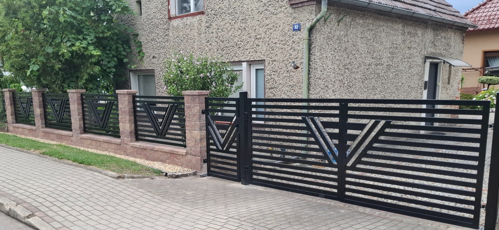 Nowoczesne, czarne ogrodzenie z poziomych paneli i bramą wjazdową, z dekoracyjnym motywem litery V, osadzone na niskim murku z cegły. Dom w tle.