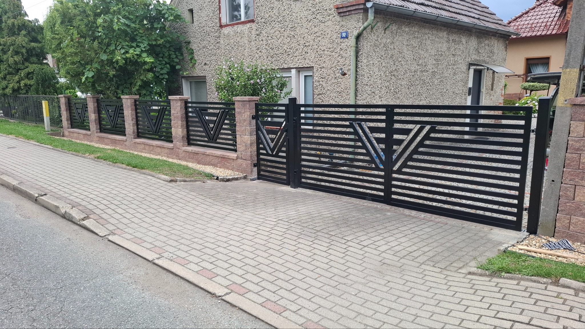 Nowoczesne ogrodzenie metalowe z bramą wjazdową, czarne poziome panele z motywem V, murowane słupki z cegły klinkierowej, kostka brukowa na podjeździe.