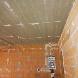 BRYLL ELECTRIC ARKADIUSZ BRYLL - Surowy stan wnętrza w trakcie prac: widoczne ceglane ściany, izolacja sufitu z poprowadzonymi przewodami oraz puszka elektryczna z kablami w rurze ochronnej.