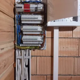 Rozdzielnica elektryczna z zabezpieczeniami i przewodami, zamontowana na ścianie z cegły dziurawki, obok ściany z drewnianych paneli.
