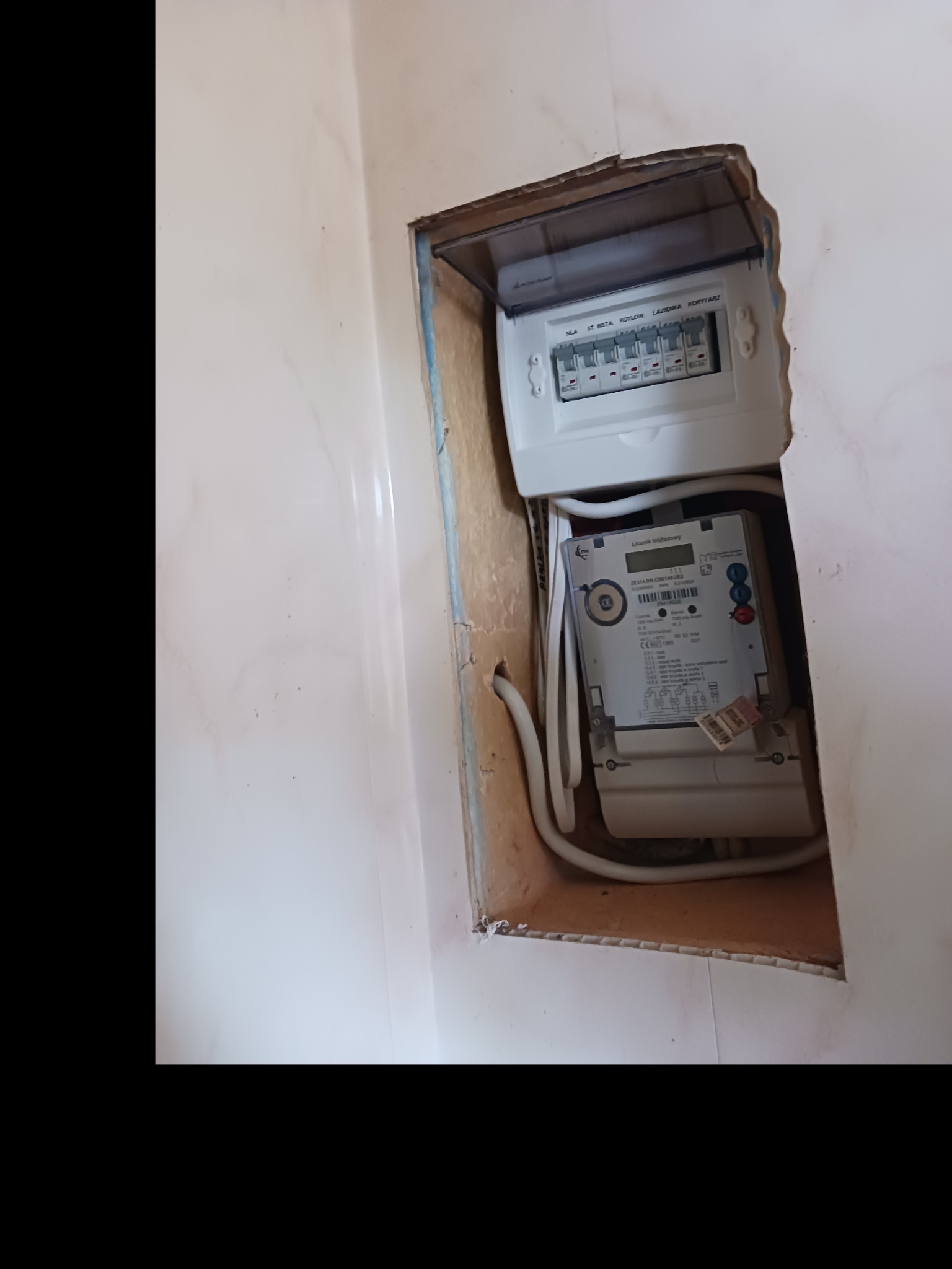Otwarta skrzynka z bezpiecznikami i zamontowany pod nią licznik elektryczny w płytkiej wnęce wyłożonej płytkami.