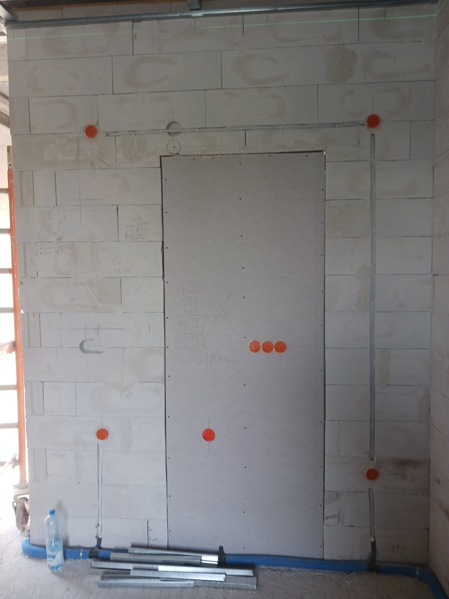 Ściana z betonu komórkowego z wywierconymi otworami pod puszki elektryczne, widoczne pionowe bruzdy pod kable i poziome profile montażowe pod sufitem, niebieski karbowany peszel i butelka wody...