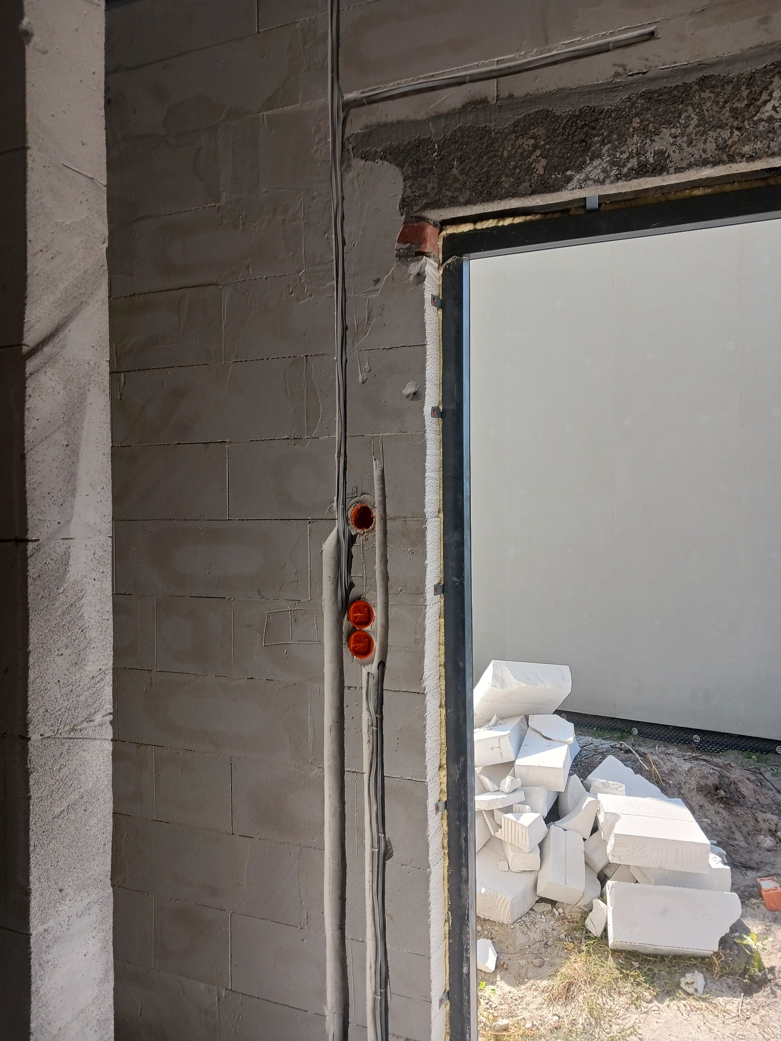 Ściana z betonu komórkowego w trakcie prac instalacyjnych, widoczne kable elektryczne w peszlach i puszki podtynkowe w kolorze pomarańczowym.