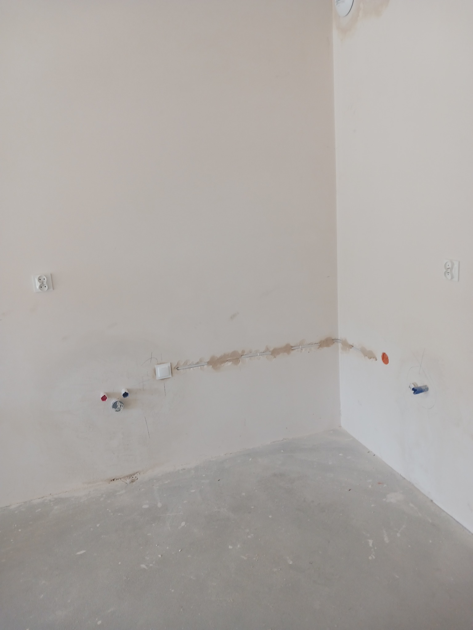 Surowe wnętrze w trakcie prac wykończeniowych: widoczne puszki elektryczne, gniazdka, świeżo położona gładź na ścianach i surowy beton na podłodze.