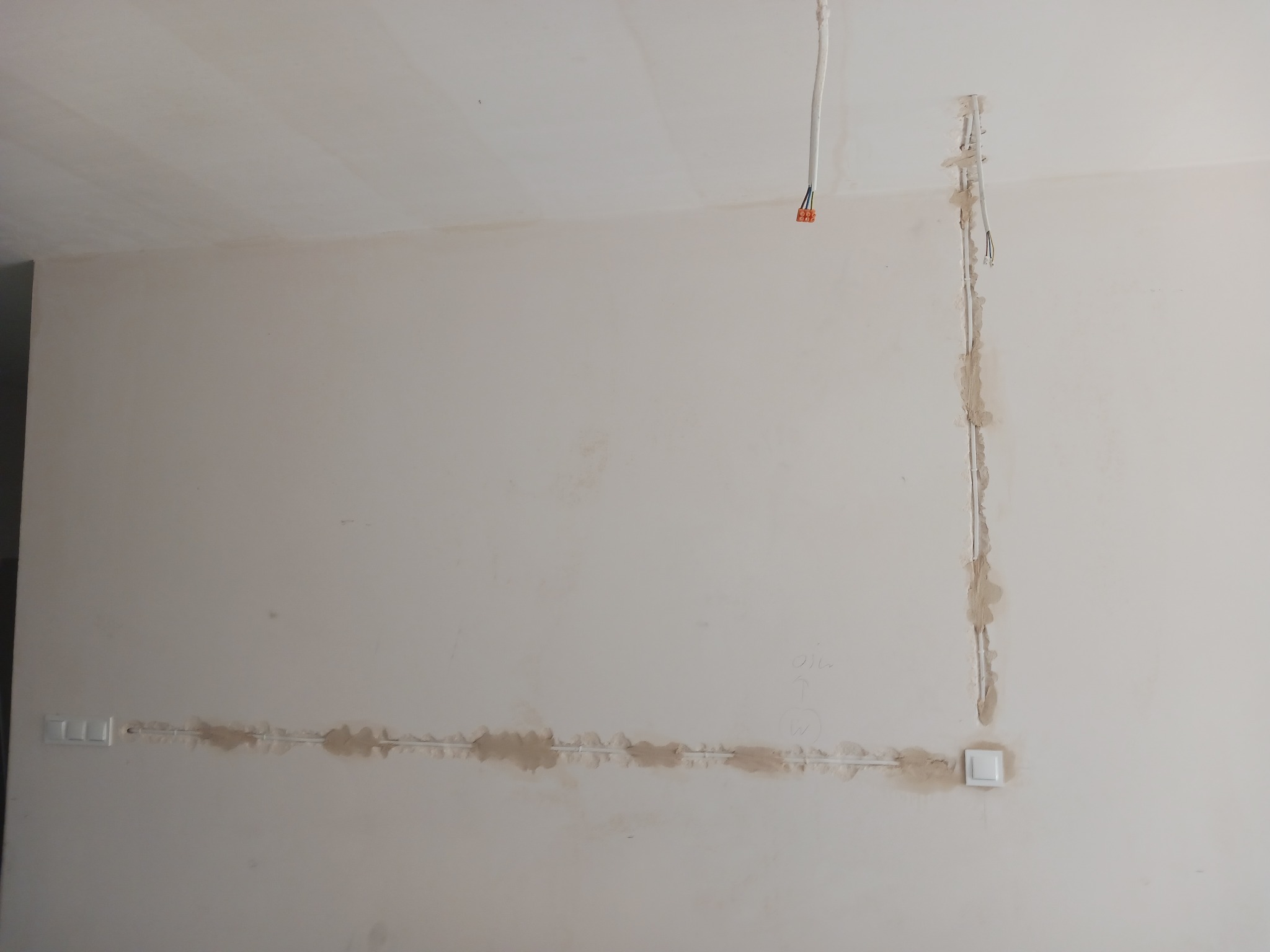 Biała ściana w trakcie remontu z widocznymi przewodami elektrycznymi w bruzdach, gniazdko i włącznik światła, przygotowane do dalszych prac wykończeniowych.