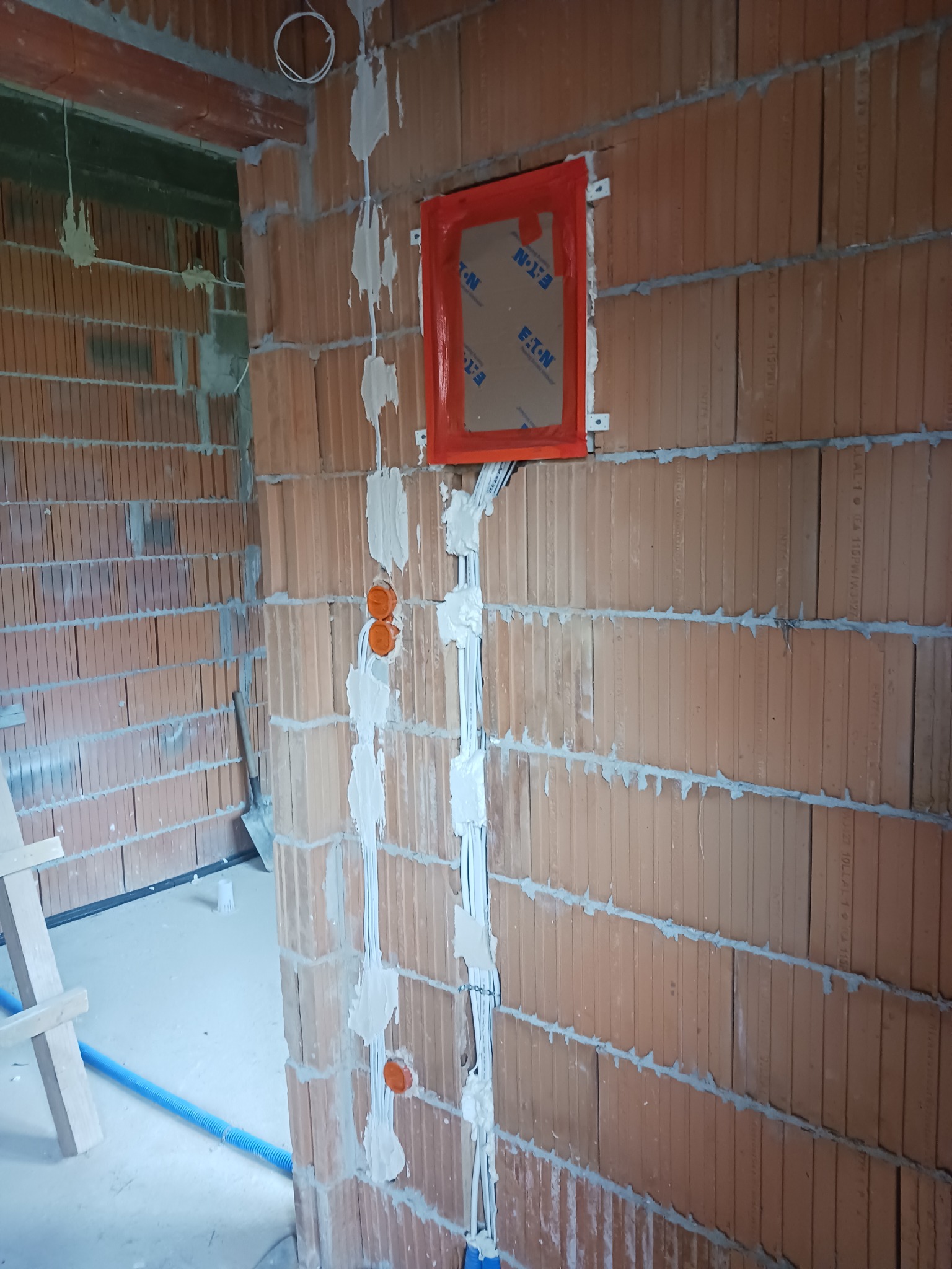 Ściana z czerwonej cegły z widocznymi przewodami elektrycznymi zabezpieczonymi białym gipsem, pomarańczowe puszki instalacyjne oraz otwór z czerwoną ramką, w tle widoczny fragment pomieszczenia...