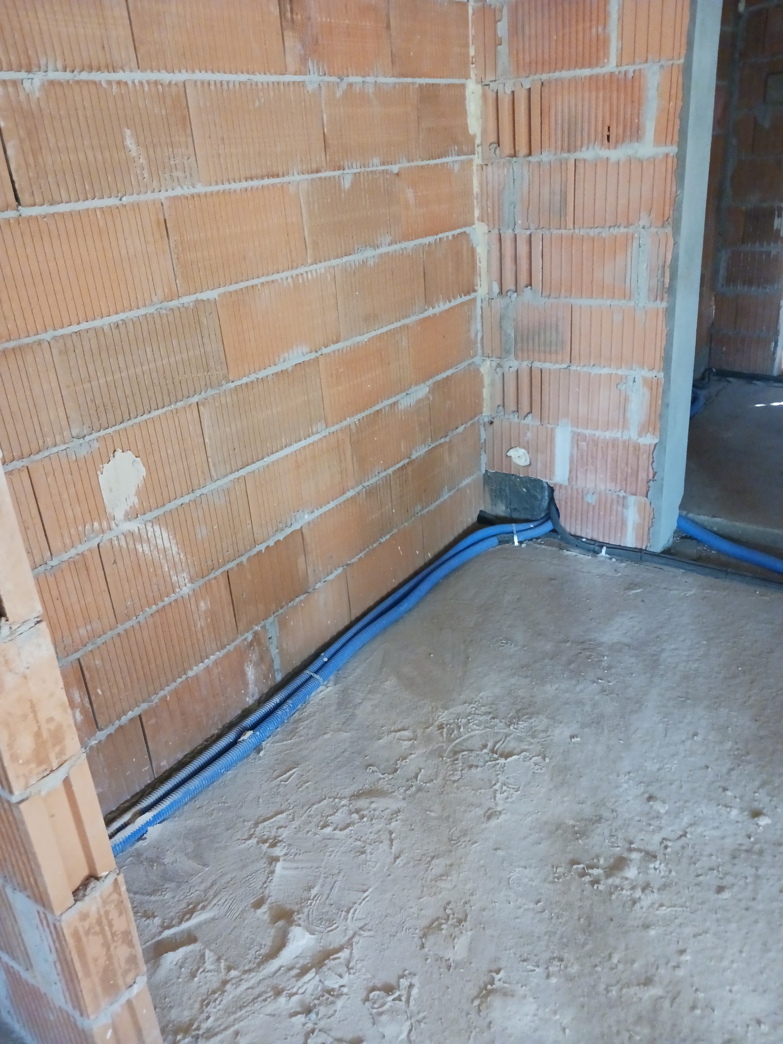 Dwa niebieskie peszle elektryczne prowadzone wzdłuż ściany z czerwonej cegły dziurawki, przymocowane do podłoża białymi uchwytami, widok na surową posadzkę.