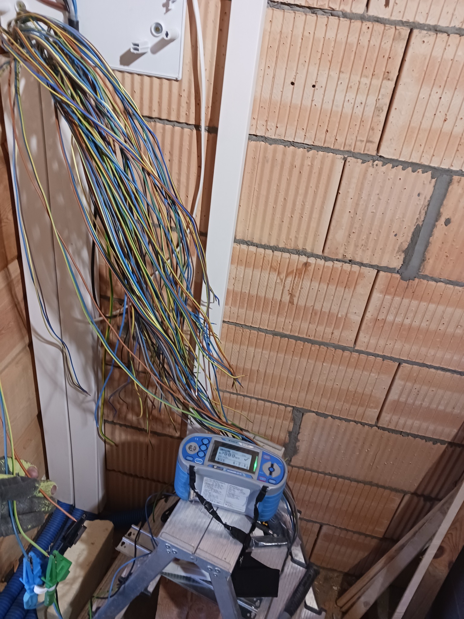 Wiązka kolorowych przewodów wychodząca ze skrzynki elektrycznej na tle surowej ściany z cegieł, miernik elektryczny na aluminiowej drabinie w dolnej części kadru.