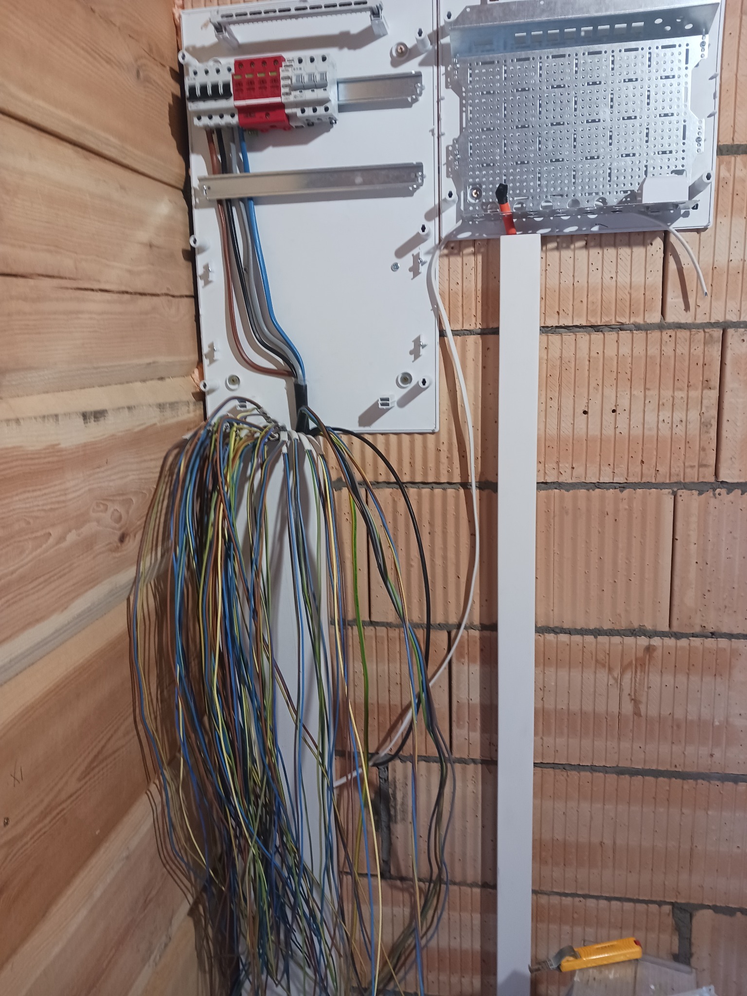 Rozdzielnica elektryczna w trakcie montażu w surowym wnętrzu, widoczne liczne przewody, biała obudowa na ścianie z cegły i drewnianej boazerii, narzędzie w dolnym rogu.