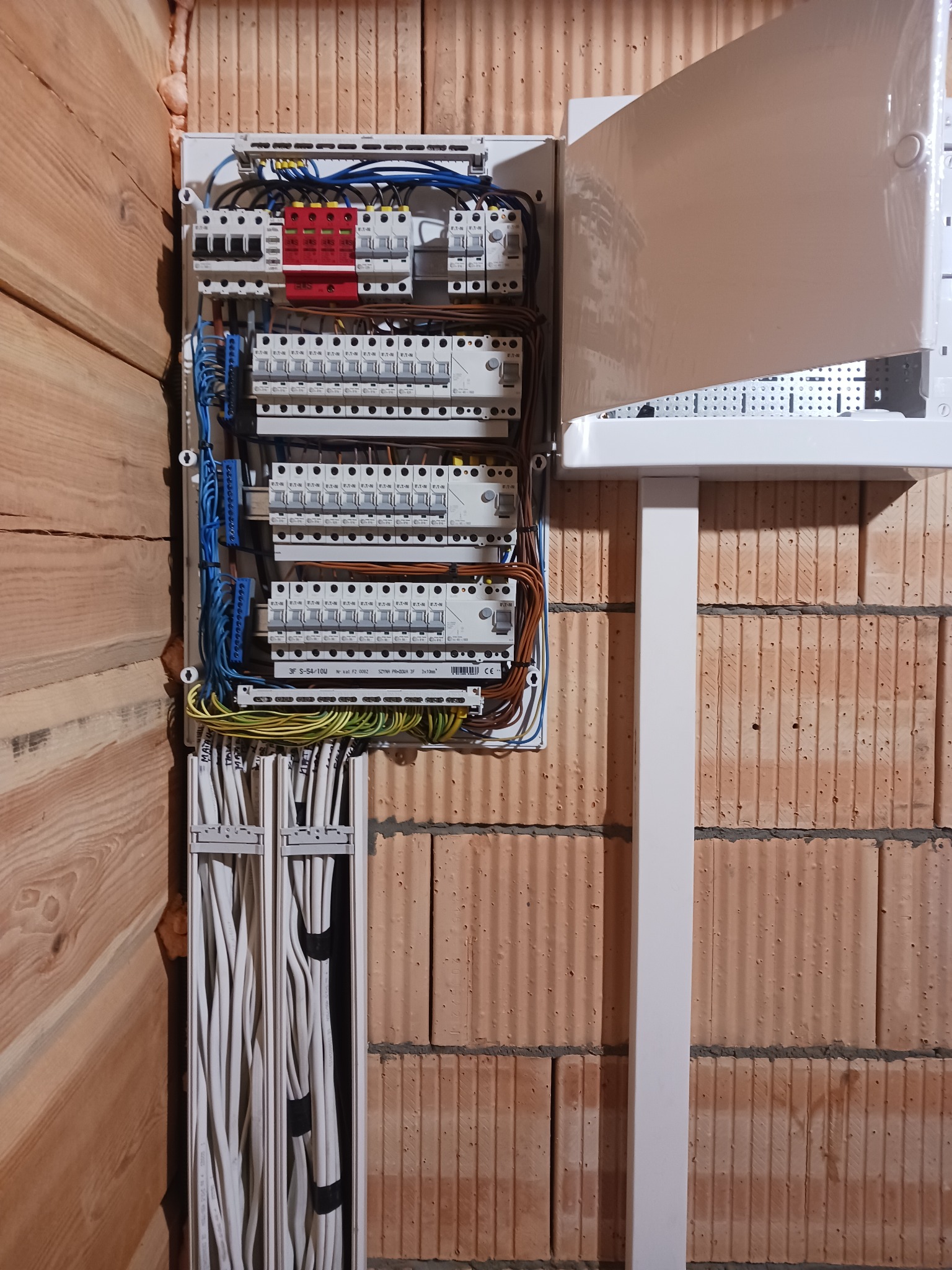 Rozdzielnica elektryczna z zabezpieczeniami i przewodami, zamontowana na ścianie z cegły dziurawki, obok ściany z drewnianych paneli.