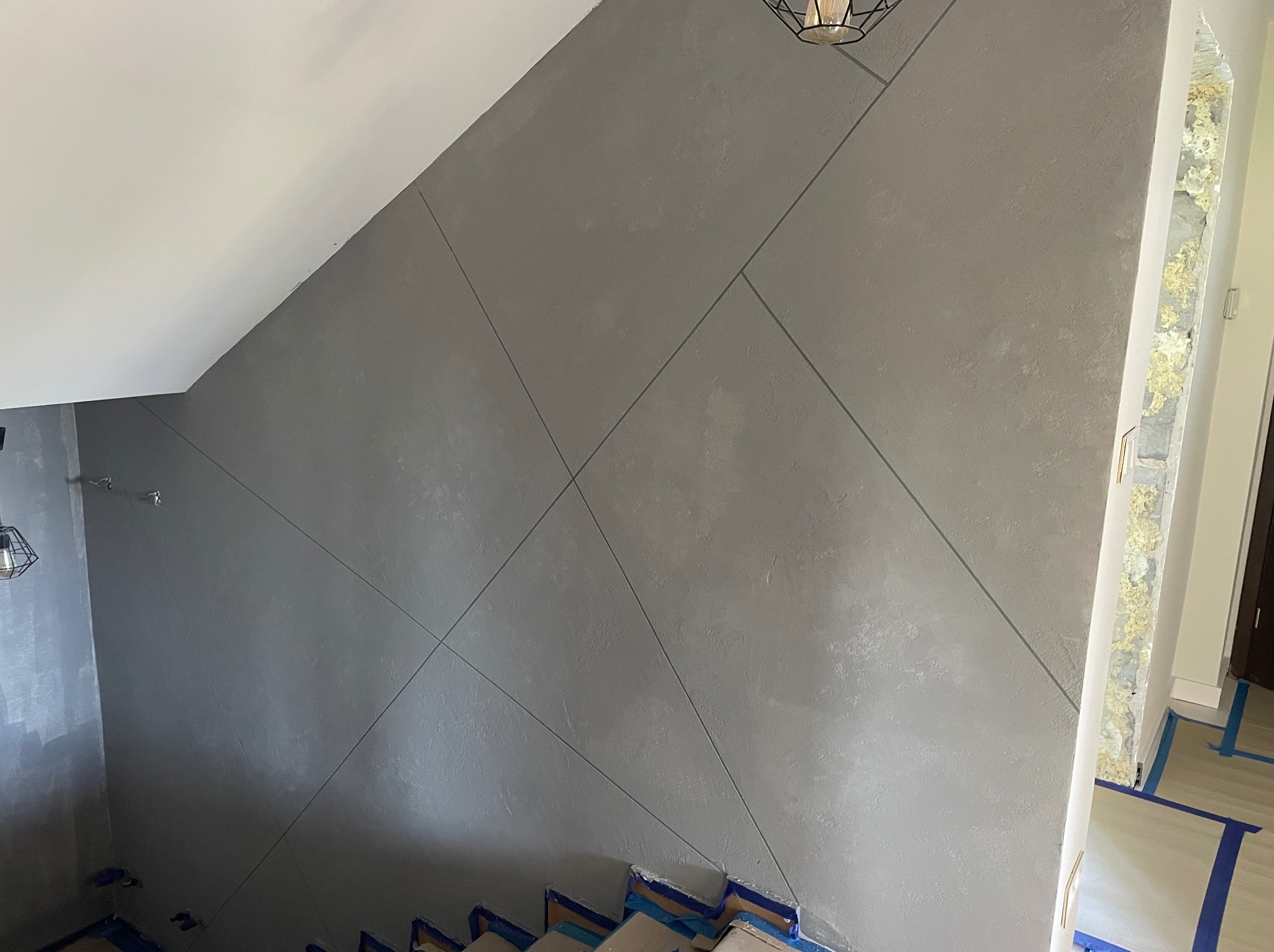 Szara ściana w klatce schodowej z dekoracyjnym wzorem geometrycznym z ciemniejszych linii, zabezpieczone schody taśmą malarską.