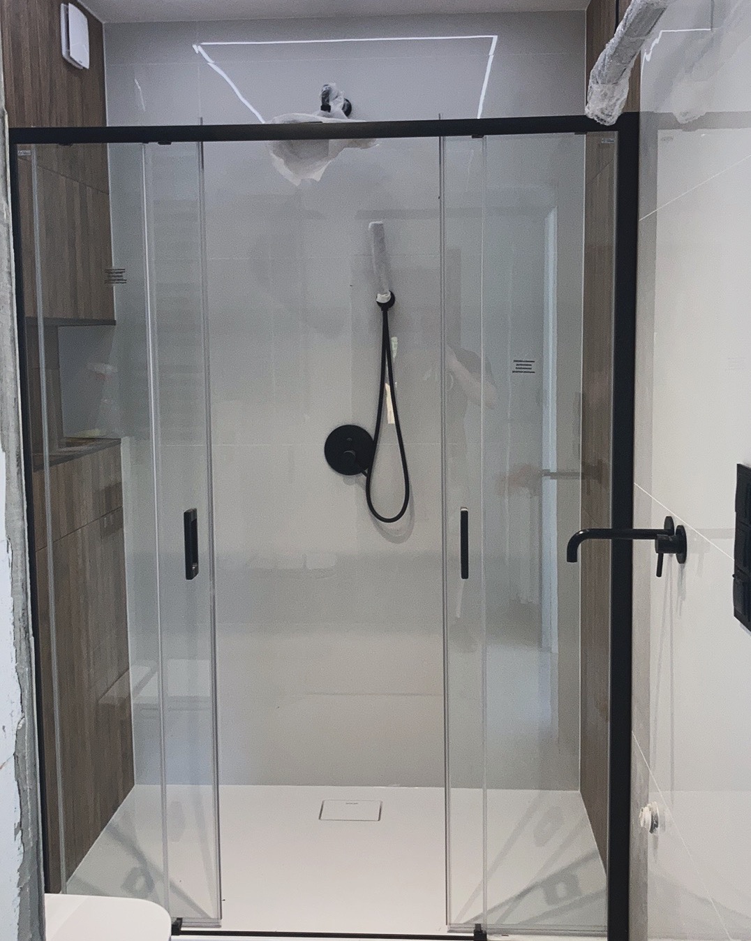 Nowoczesna kabina prysznicowa z czarnymi profilami i armaturą, częściowo zabezpieczona folią ochronną, widoczne białe płytki i drewnopodobna okładzina ścienna.