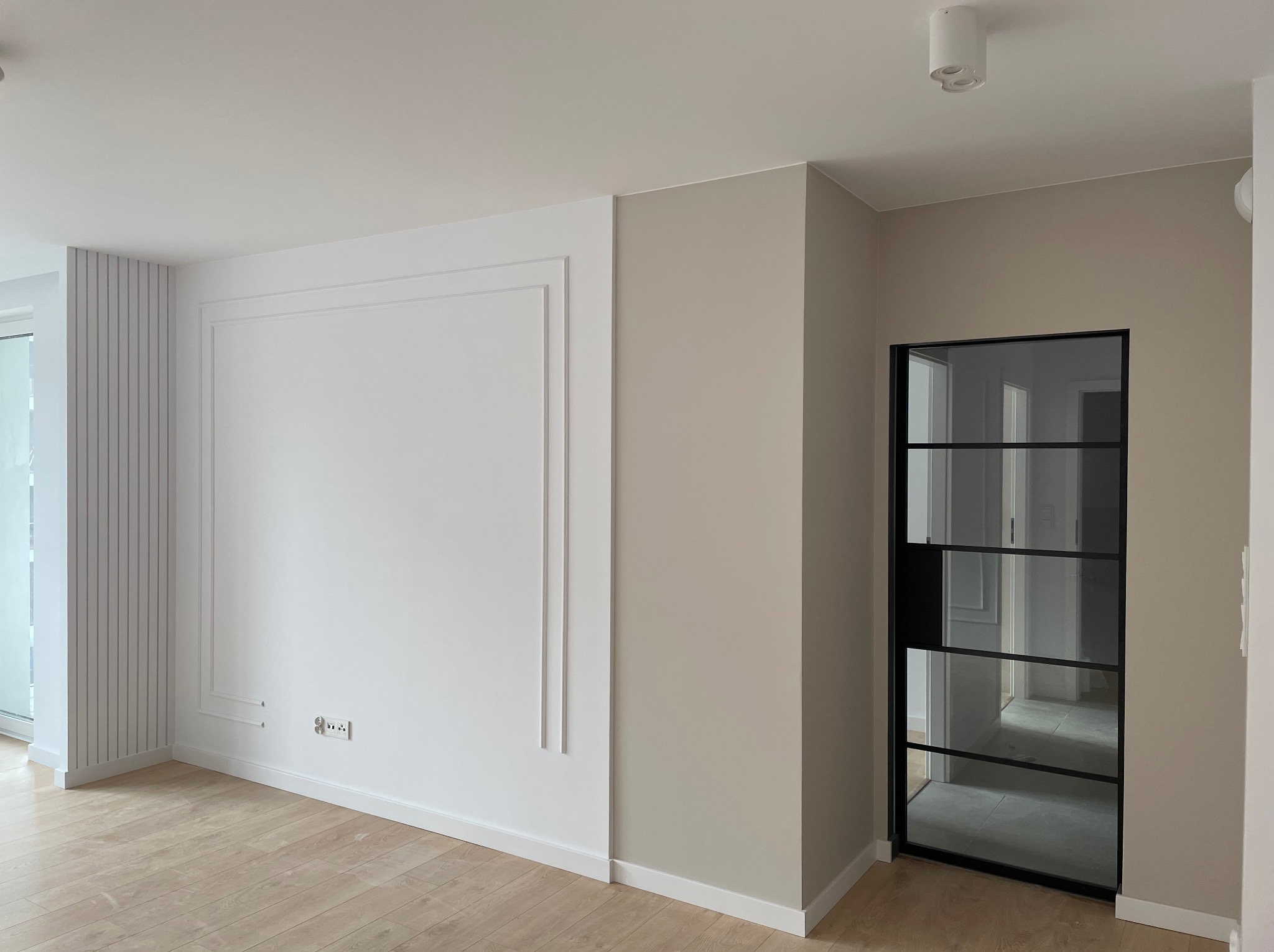 Wnętrze po remoncie: biała ściana z dekoracyjnymi listwami i ramą, jasny parkiet, ściana w odcieniach beżu, czarne drzwi szklane z podziałami