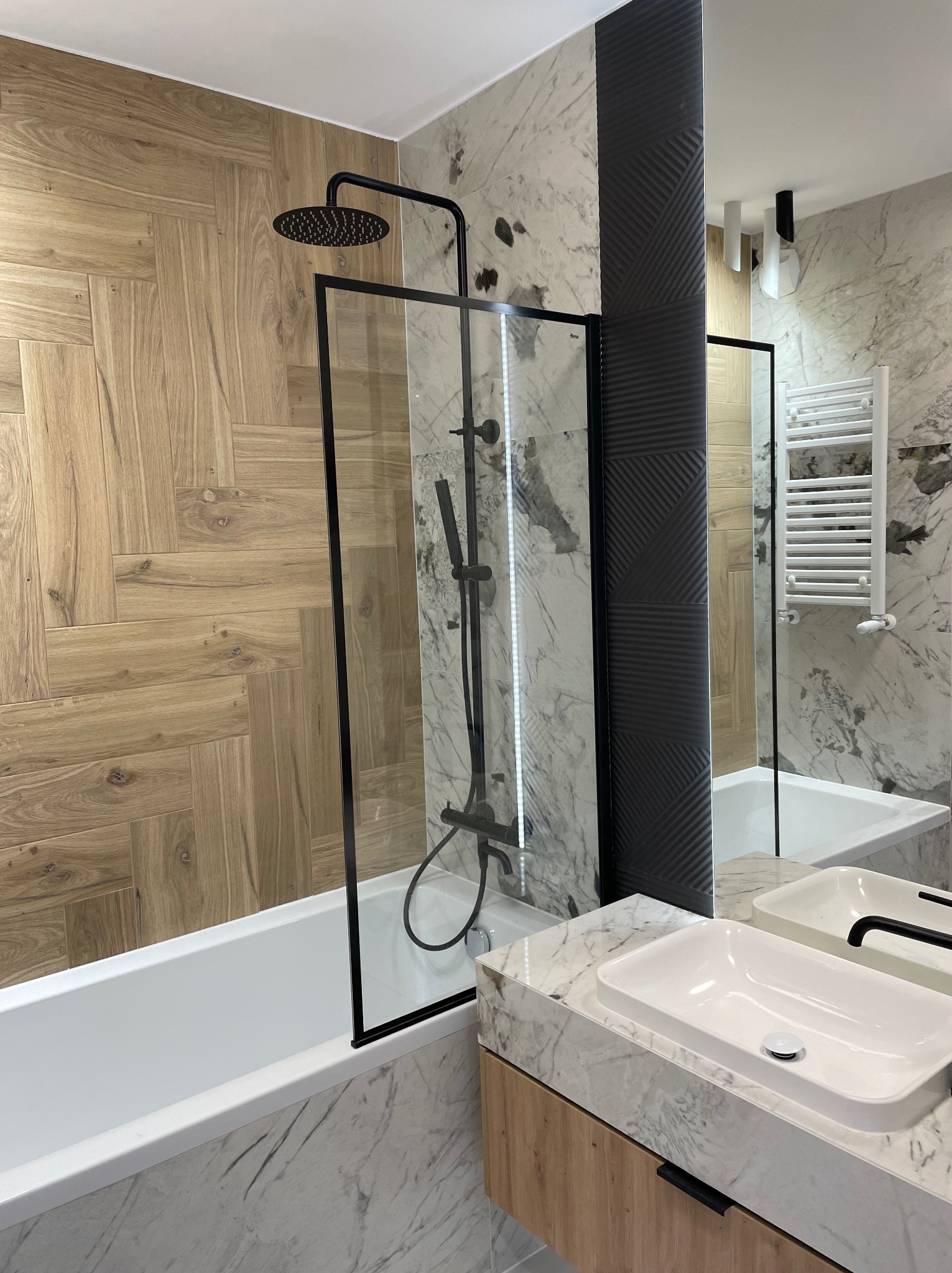 Nowoczesna łazienka z wanną z parawanem, deszczownicą w kolorze czarnym, umywalką nablatową na blacie z imitacji marmuru oraz drewnopodobnymi płytkami ułożonymi w jodełkę na ścianie.