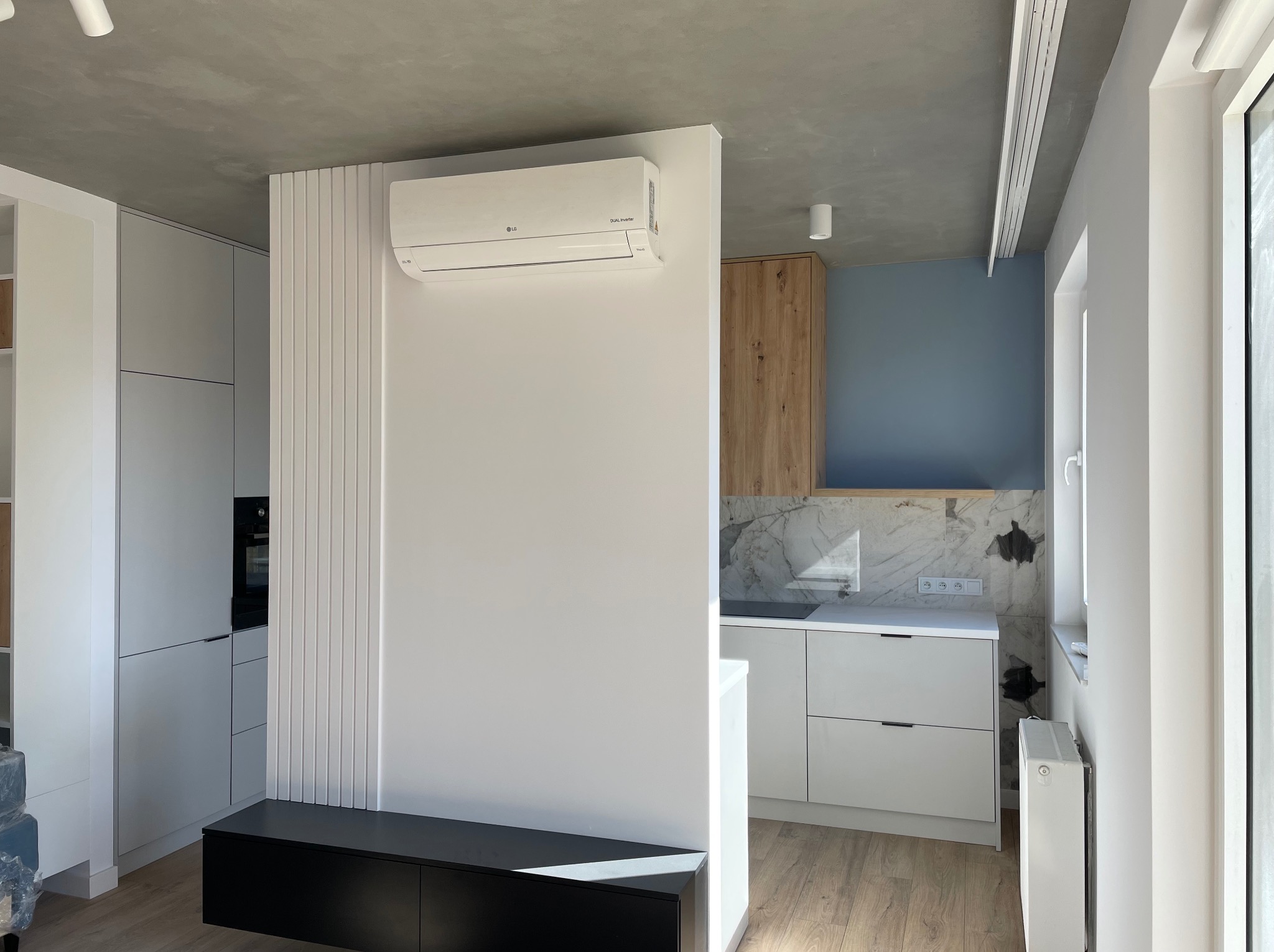 Nowoczesne wnętrze: biała ściana z klimatyzatorem LG, dekoracyjna ścianka z pionowych listew, czarna szafka RTV, fragment kuchni z białymi szafkami i marmurowym backsplash'em, szary sufit.