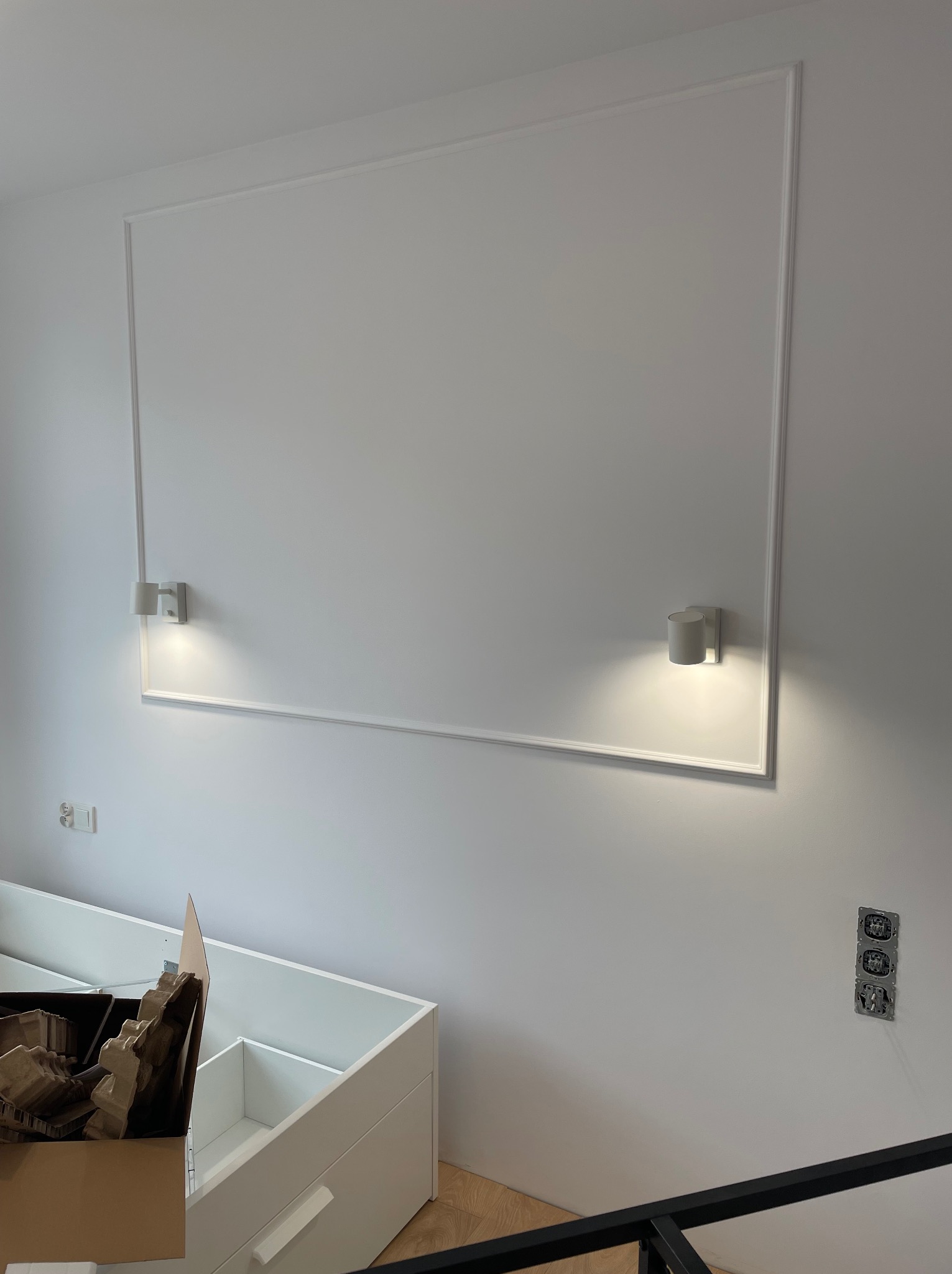 Biała ściana z dekoracyjną ramą, dwoma kinkietami i niezamontowanymi gniazdkami elektrycznymi; w dolnej części kadru widoczne elementy białych mebli w trakcie montażu.