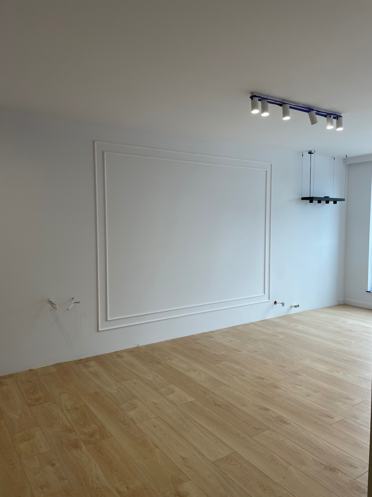 Pusty pokój po remoncie z panelami podłogowymi, białymi ścianami z dekoracyjną ramą, nowoczesnym oświetleniem sufitowym i wystającymi przewodami elektrycznymi.