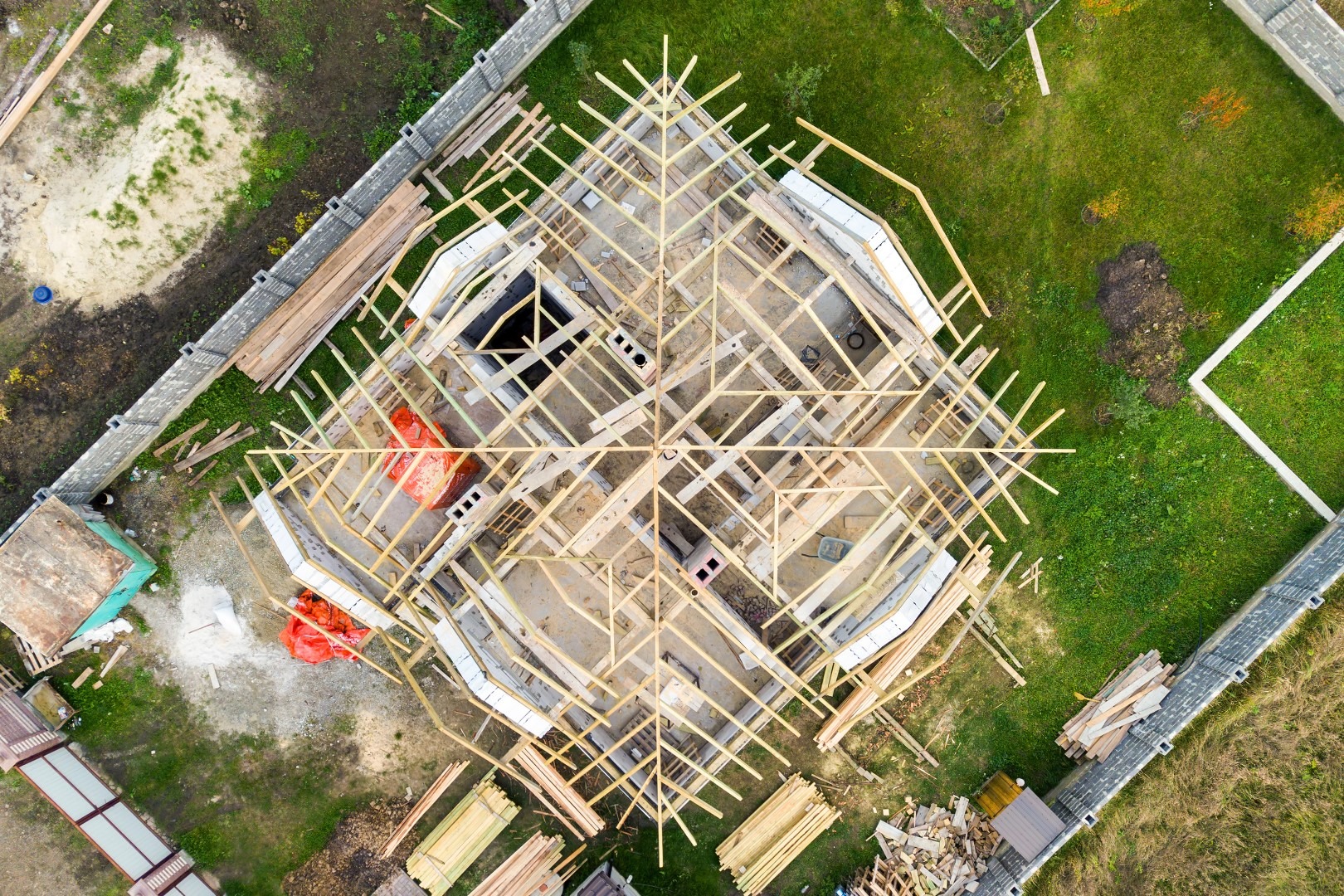Widok z góry na budowę domu jednorodzinnego w trakcie montażu drewnianej konstrukcji dachu, z widocznymi elementami fundamentów i materiałami budowlanymi na działce.