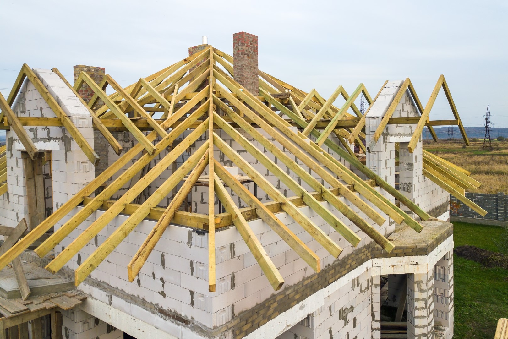 Surowa konstrukcja domu z widoczną więźbą dachową i kominami z cegły, w tle pola i słupy wysokiego napięcia.