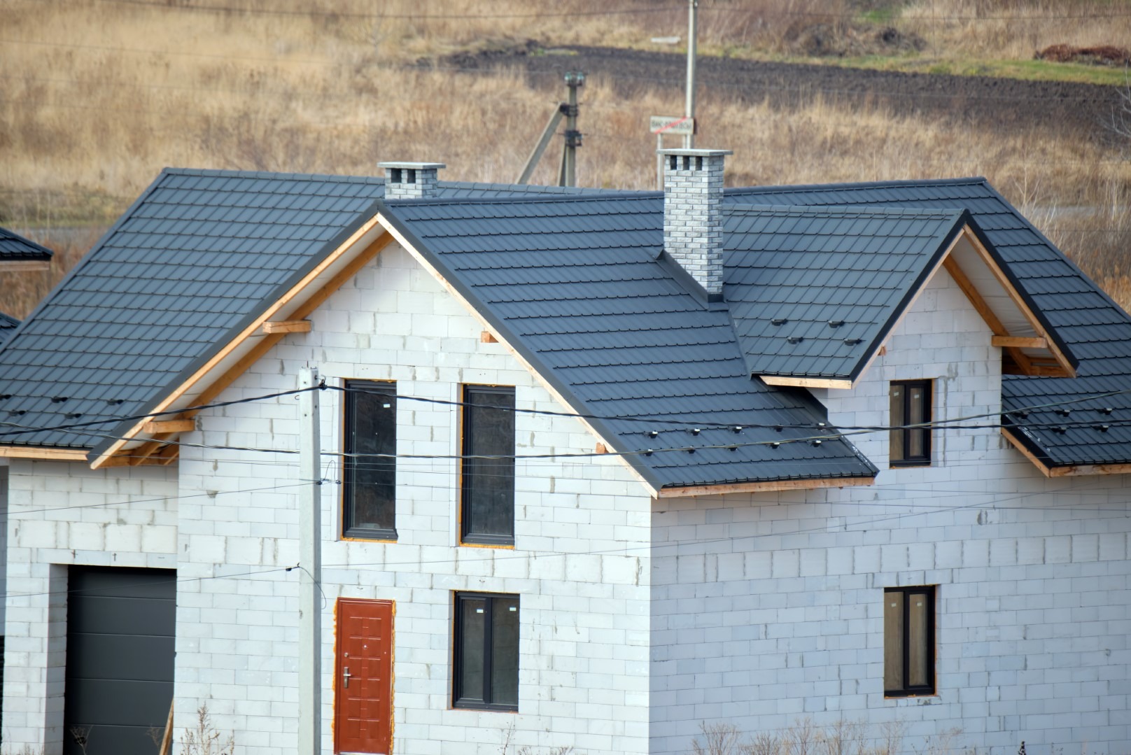 Niedokończony dom w stanie surowym zamkniętym z ciemnoszarą dachówką, widoczne kominy z cegły i pustaków, drewniana więźba dachowa, brązowe drzwi wejściowe.