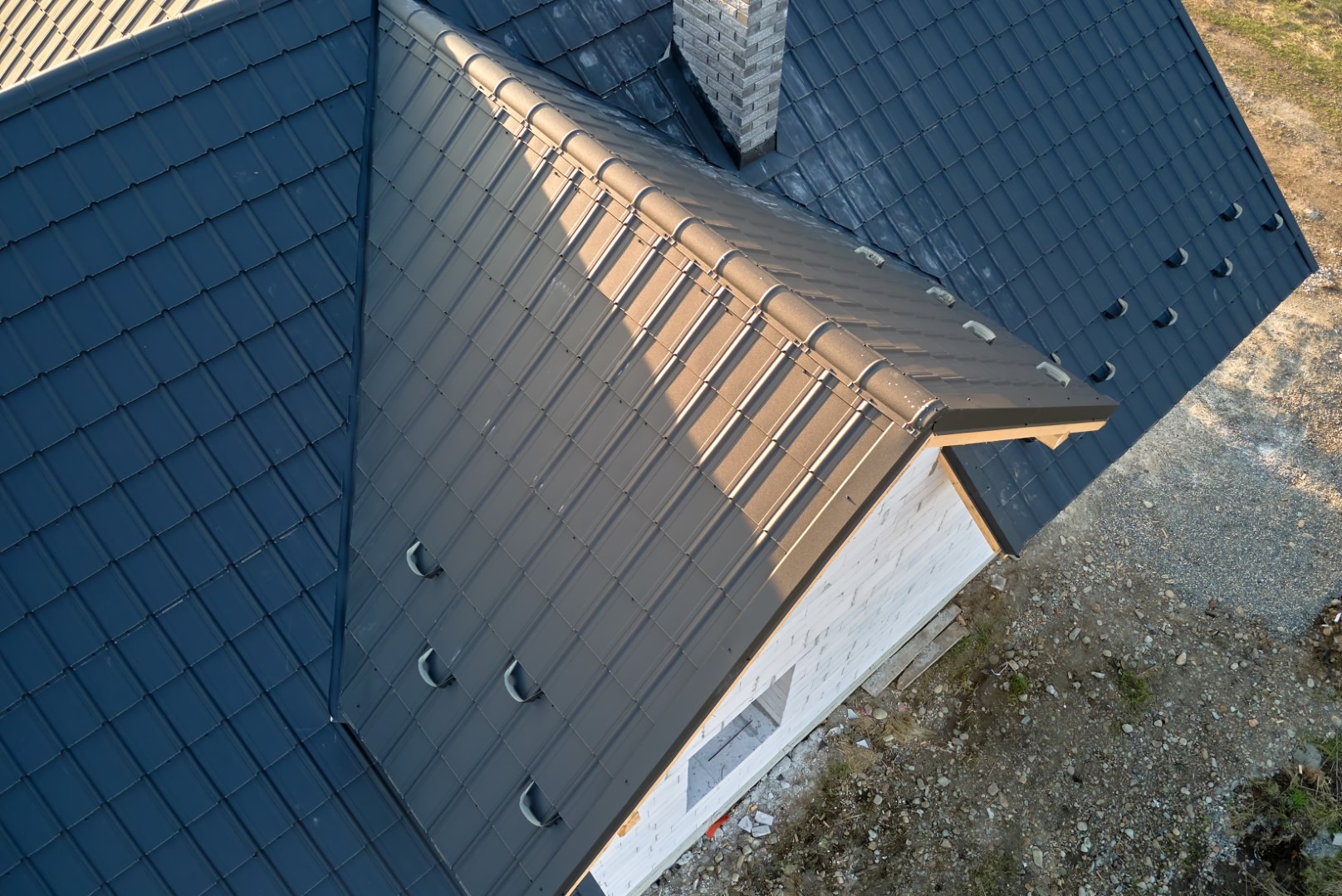 Widok z góry na nowo położony, dwuspadowy dach z ciemnej dachówki ceramicznej i jasnoszarej blachy trapezowej na domu w trakcie budowy, z kominem z cegły klinkierowej.