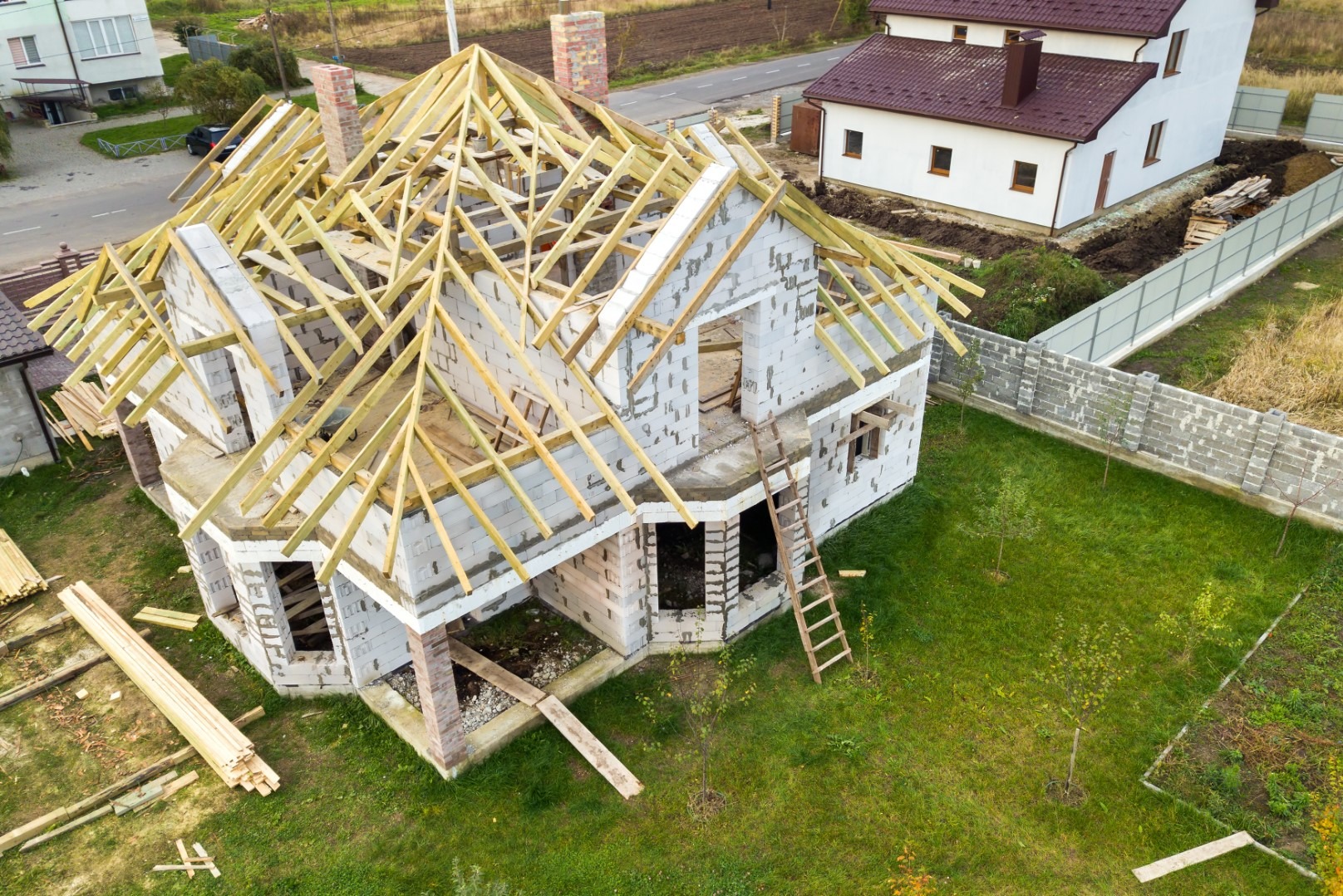 Budowa domu jednorodzinnego z widoczną konstrukcją drewnianą dachu, ściany z białych bloczków, drabina oparta o budynek, widok z góry na posesję z trawnikiem i ogrodzeniem.
