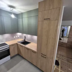 Nowoczesna kuchnia z drewnopodobnymi frontami, czarnym zlewem i szarą zabudową ścienną z pionowymi panelami. W tle widoczne odbicie fotografa w lustrzanej szafie.