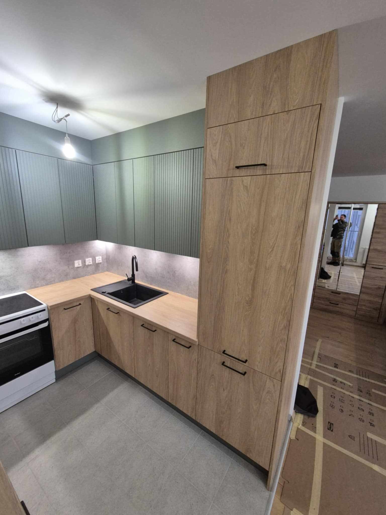 Nowoczesna kuchnia z drewnopodobnymi frontami, czarnym zlewem i szarą zabudową ścienną z pionowymi panelami. W tle widoczne odbicie fotografa w lustrzanej szafie.