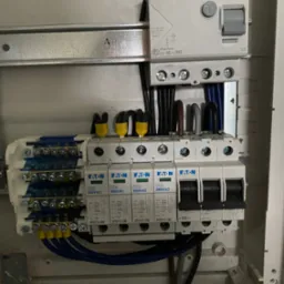 Szczegółowe ujęcie wnętrza skrzynki elektrycznej z zainstalowanymi bezpiecznikami EATON, przewodami z żółtymi końcówkami i wyłącznikiem różnicowoprądowym, widoczne oznaczenia L1, L2, L3, N.