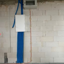 EL-KOS Usługi Elektryczne - Niebieskie rury i pomarańczowe przewody elektryczne na ścianie z betonu komórkowego w trakcie instalacji w Zielinie. Widoczna skrzynka rozdzielcza.