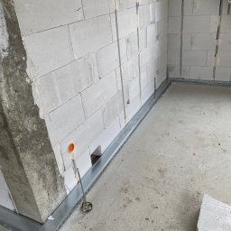 EL-KOS Usługi Elektryczne - Surowy stan: ściany z betonu komórkowego z puszkami elektrycznymi i metalowe korytka kablowe przy podłodze, przygotowane do dalszych prac instalacyjnych.
