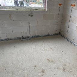 EL-KOS Usługi Elektryczne - Surowy stan: wnętrze z widoczną, częściowo zamontowaną instalacją elektryczną w postaci metalowych korytek przy ścianach i przewodów wychodzących z puszek. W tle okno z widocznym autem.