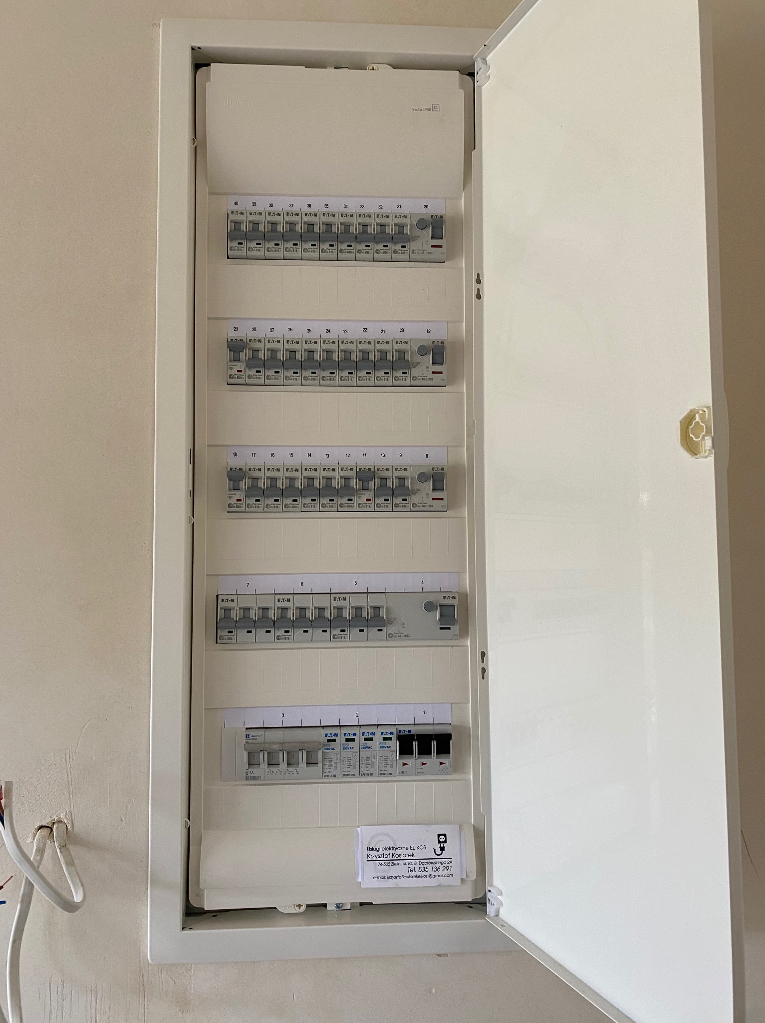 Otwarta skrzynka elektryczna z bezpiecznikami Eaton, zamontowana na ścianie, widoczne wystające kable, etykieta firmy elektrycznej EL-KOS wewnątrz.