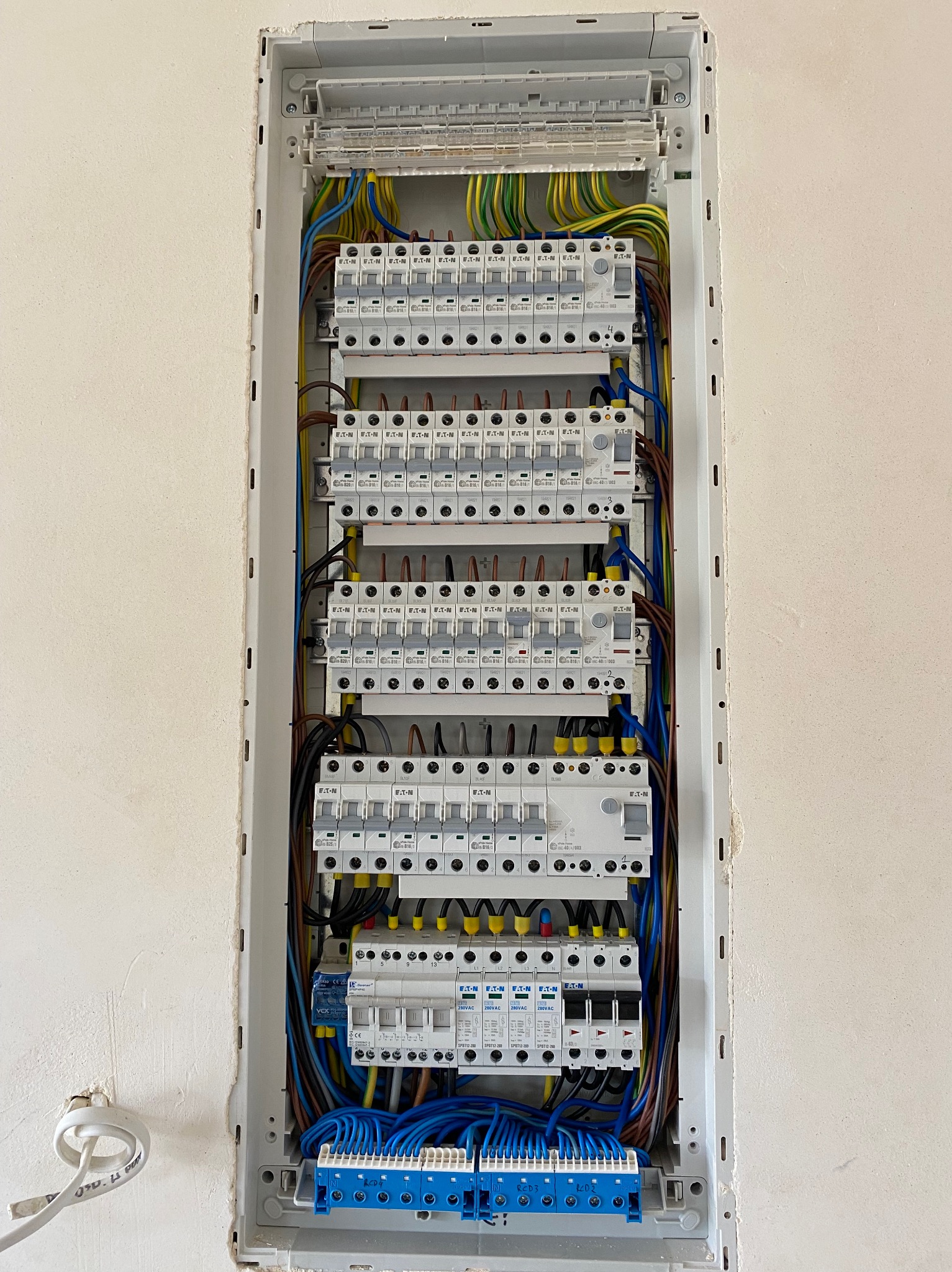 Otwarta skrzynka rozdzielcza z zainstalowanymi bezpiecznikami i okablowaniem w kolorach niebieskim, żółto-zielonym i brązowym, zamontowana w ścianie w surowym stanie.