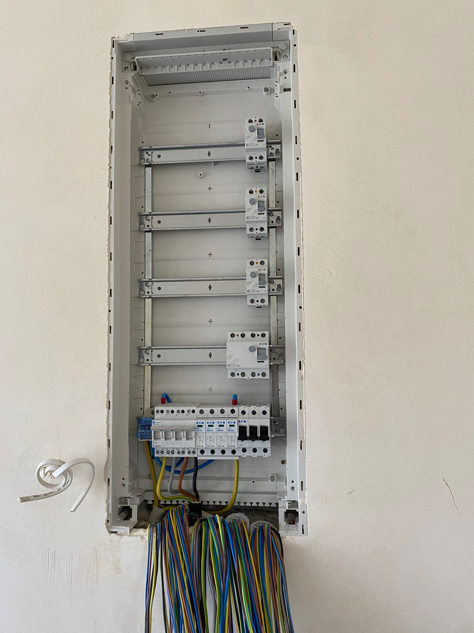 Wnęka w surowej ścianie z zamontowaną, częściowo okablowaną rozdzielnicą elektryczną. Widoczne zabezpieczenia nadprądowe Eaton i przewody w różnych kolorach.