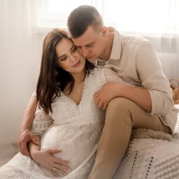Grażyna Bentkowska Fotografia - Czułe ujęcie przyszłej mamy w koronkowej sukience, obejmowanej przez partnera na tle jasnego wnętrza z dekoracjami boho.