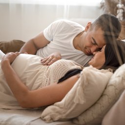 Grażyna Bentkowska Fotografia - Czułe ujęcie pary: przyszła mama w zwiewnej sukience leży na łóżku, a partner delikatnie dotyka jej brzucha, twarze blisko w intymnym geście.