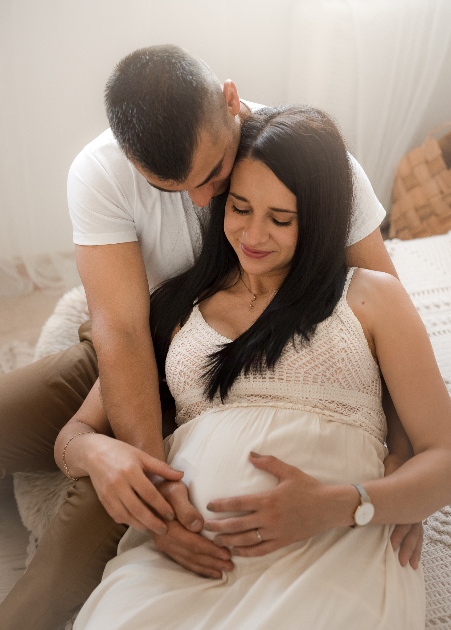 Przyszła mama w jasnej sukience, siedzi na łóżku, obejmowana przez partnera, oboje trzymają ręce na brzuchu, delikatne światło.