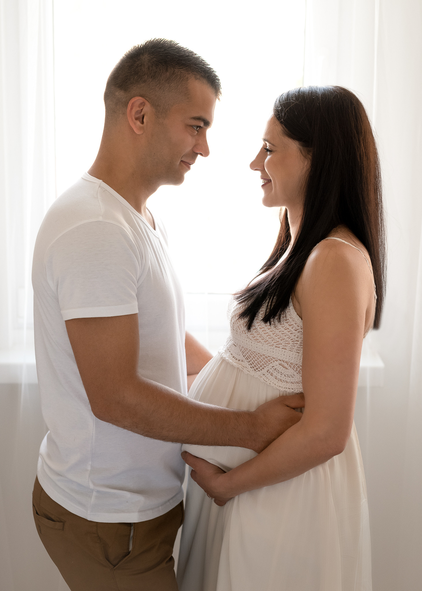 Przyszła mama w białej sukience, z ciemnymi włosami, uśmiecha się do mężczyzny, który obejmuje jej ciążowy brzuszek na tle okna z białymi zasłonami.