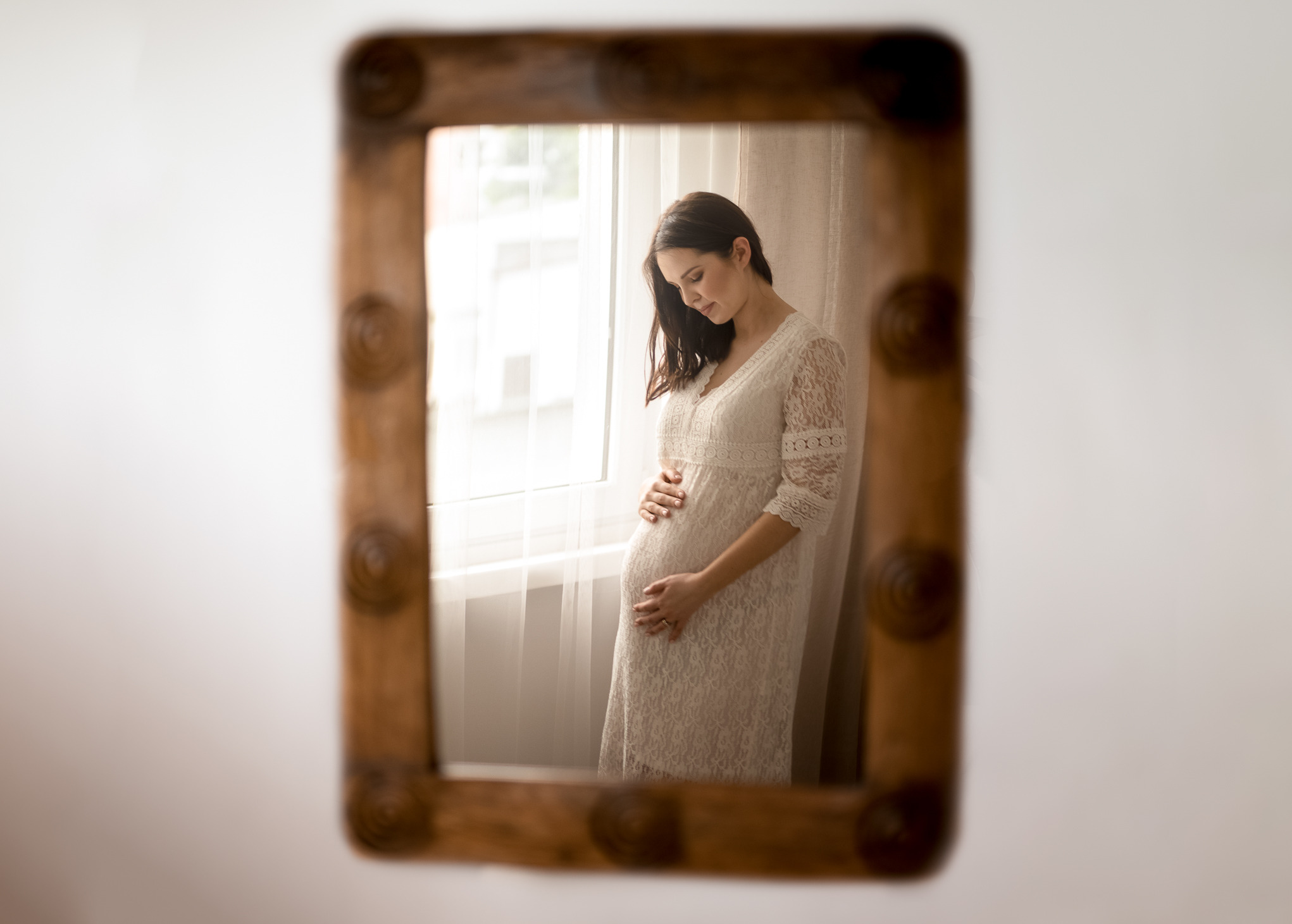 Portret kobiety w ciąży w koronkowej sukience, widziana w lustrze z drewnianą ramą, oświetlona naturalnym światłem z okna.
