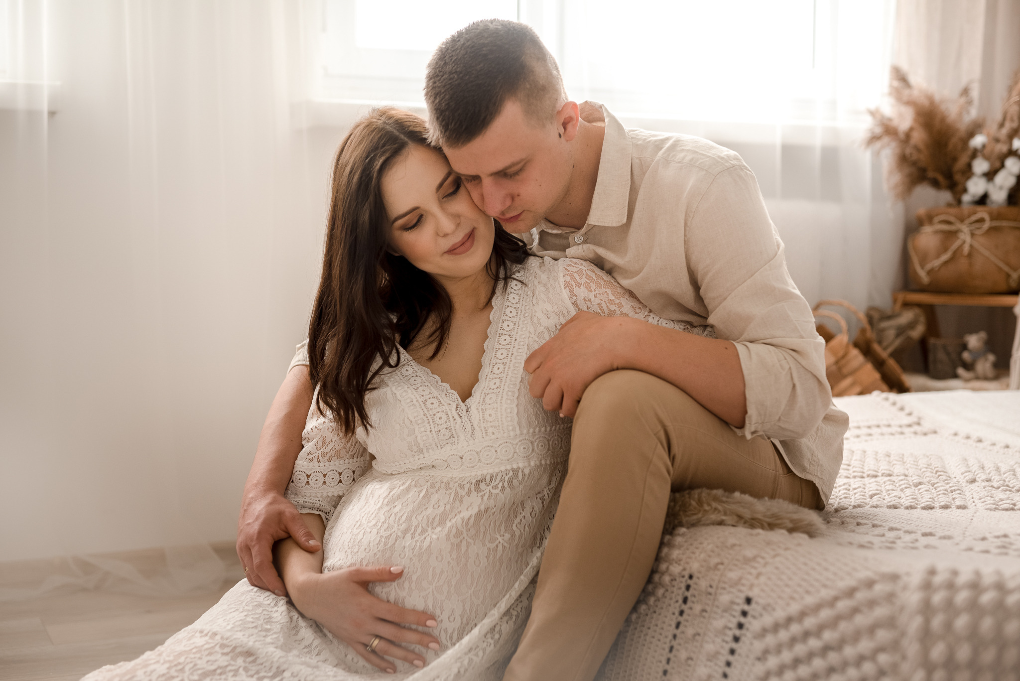 Czułe ujęcie przyszłej mamy w koronkowej sukience, obejmowanej przez partnera na tle jasnego wnętrza z dekoracjami boho.