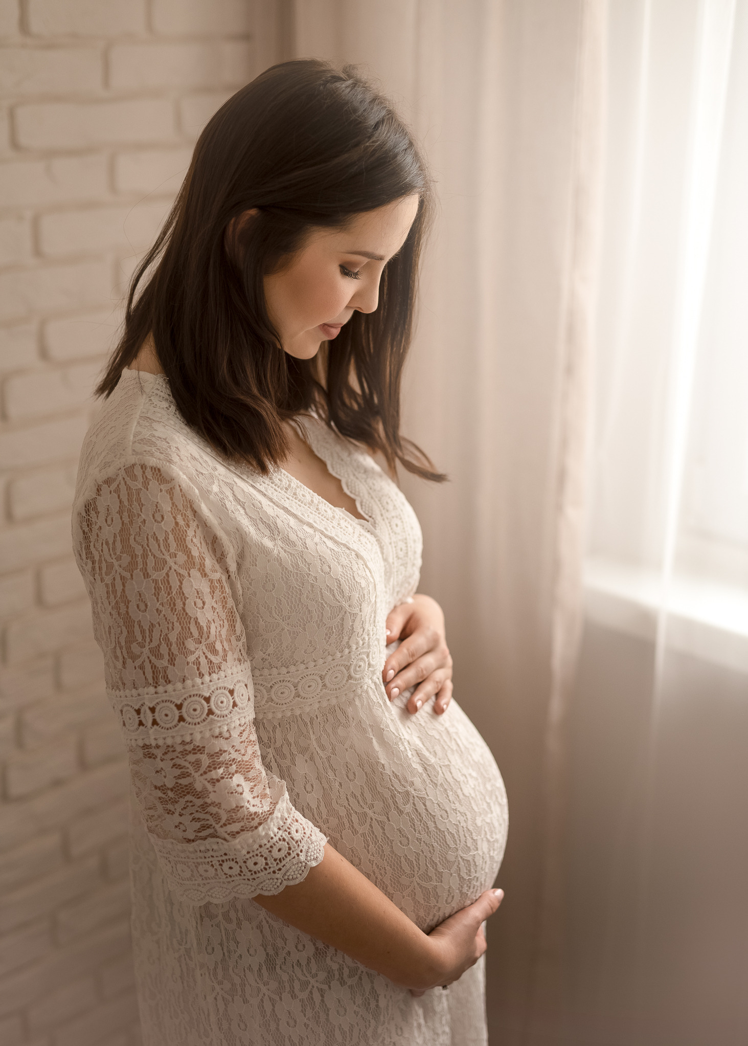 Kobieta w białej, koronkowej sukience ciążowej, z dłońmi na brzuchu, spogląda w dół w jasnym, neutralnym wnętrzu.