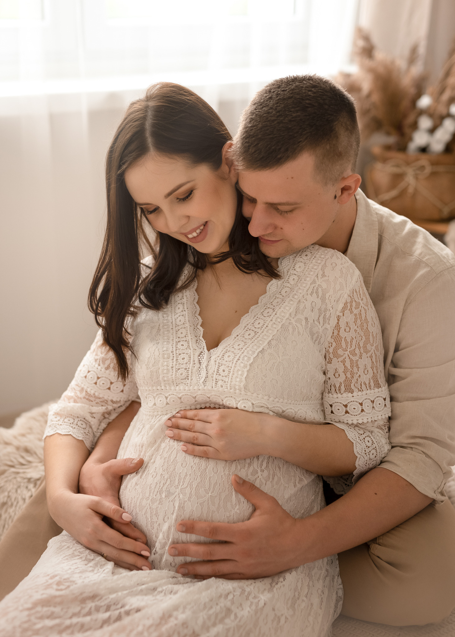 Uśmiechnięta przyszła mama w koronkowej sukience, obejmowana przez partnera, oboje trzymają ręce na brzuchu, ciepłe, naturalne światło.